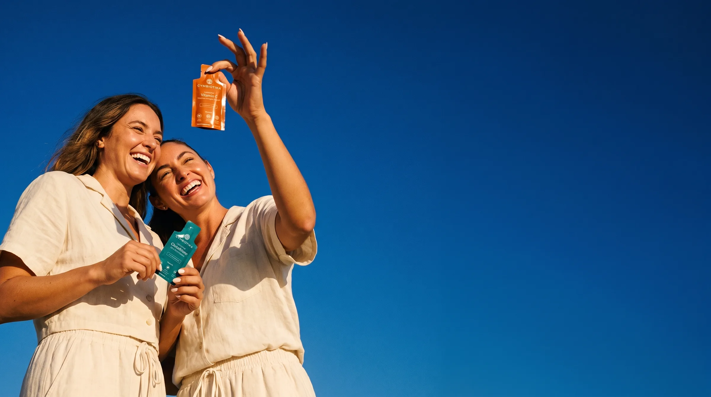 Two women laugh under a deep blue sky, one raising a small Cymbiotika Liposomal Vitamin C pouch, the other holding Liposomal Glutathione