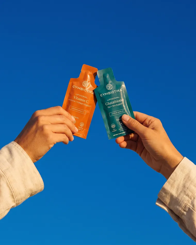 Two hands raise Cymbiotika Vitamin C and Glutathione pouches against a blue sky